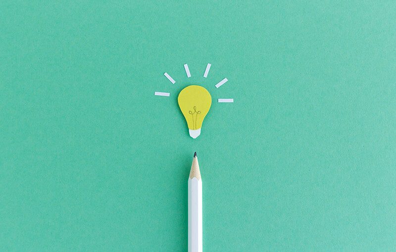 A white pencil, pointing to the top of the photo, resting on a seafoam green background. At the tip of the pencil is a paper light bulb cutout.