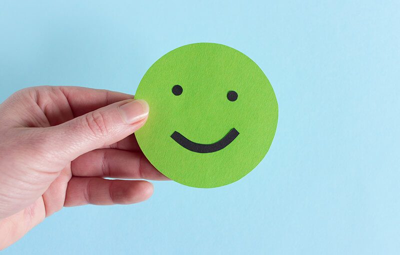 A hand holding a green paper circle with a happy face drawn on it.