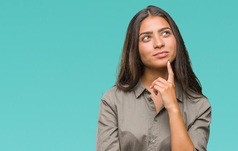 Young woman against a teal background. She has one finger pressed to her chin in a thoughtful pose.h