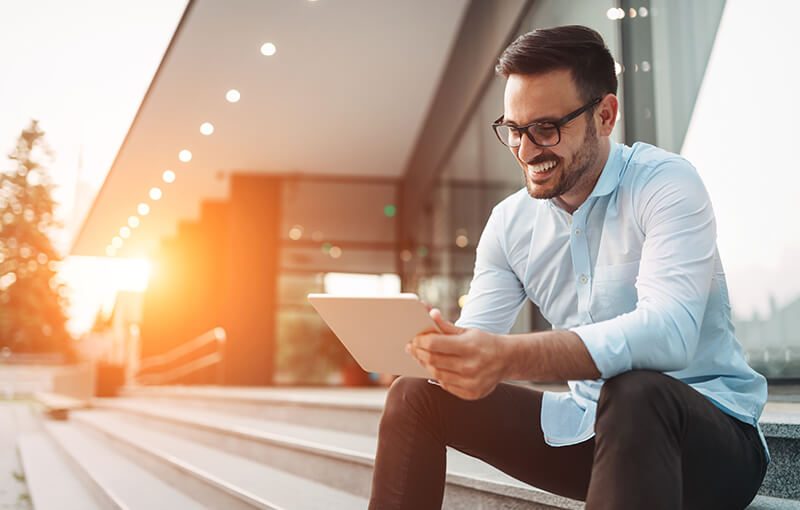 A person with short brown hair and glasses sits on the edge of some stairs in front of a large corporate building. They're smiling while they hold a tablet.