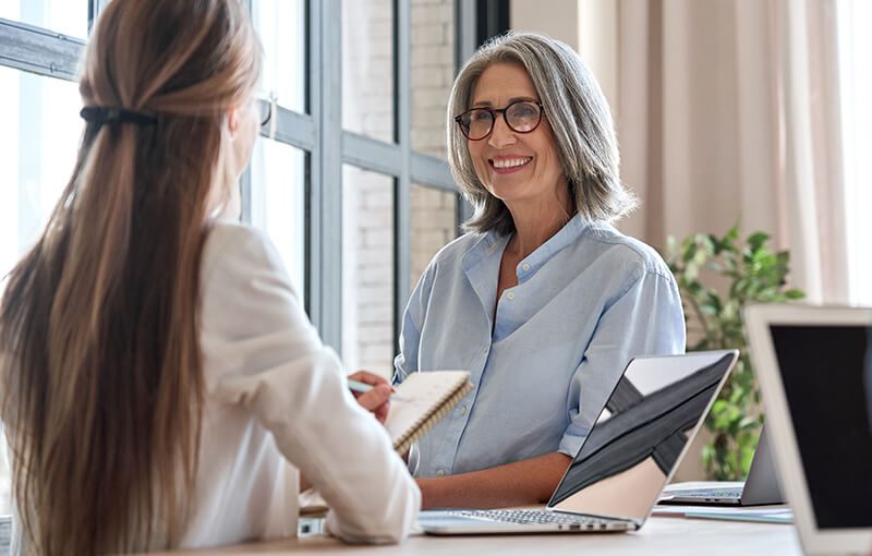 Mature, grey-haird woman smiling at a younger, long brown-haired colleague at a desk with an open laptop.