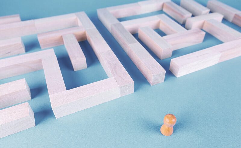 Wooden game pawn standing in front of a wooden maze on a pale, blue-grey table. The maze has a straight path through it.