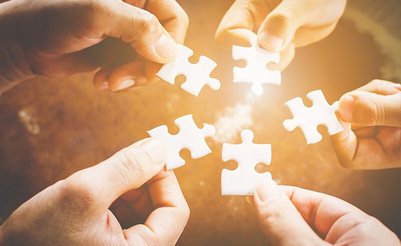 Five hands in a circle, each one holding a puzzle piece; all hands are bringing their puzzle piece to the center to combine.