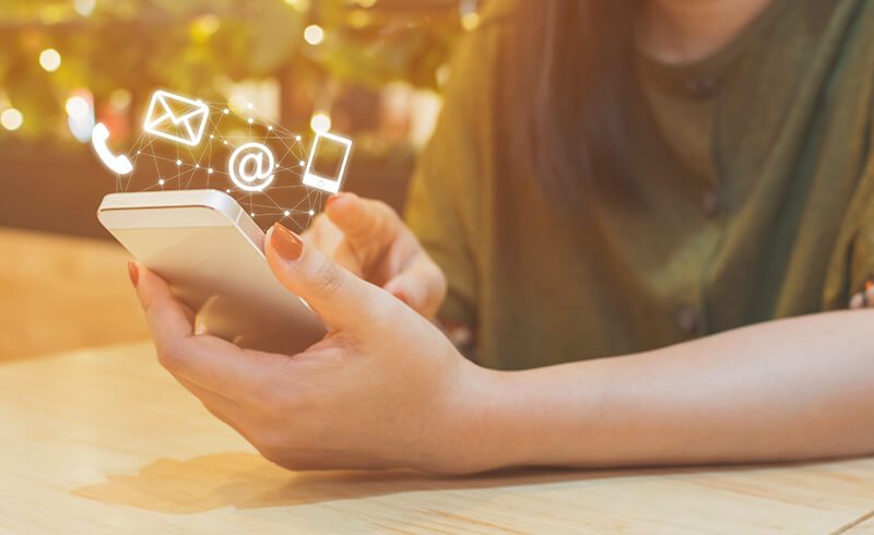 Close up on a woman's hands holding a smartphone. Above the smartphone hover generic symbols in white: an envelop, an "at" sign, and a small phone..
