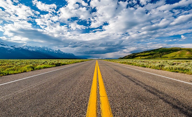 A blue and partly-cloudy sky with green fields and mountains. A paved road going in a straight line, off into the horizon line, splits the field in two.