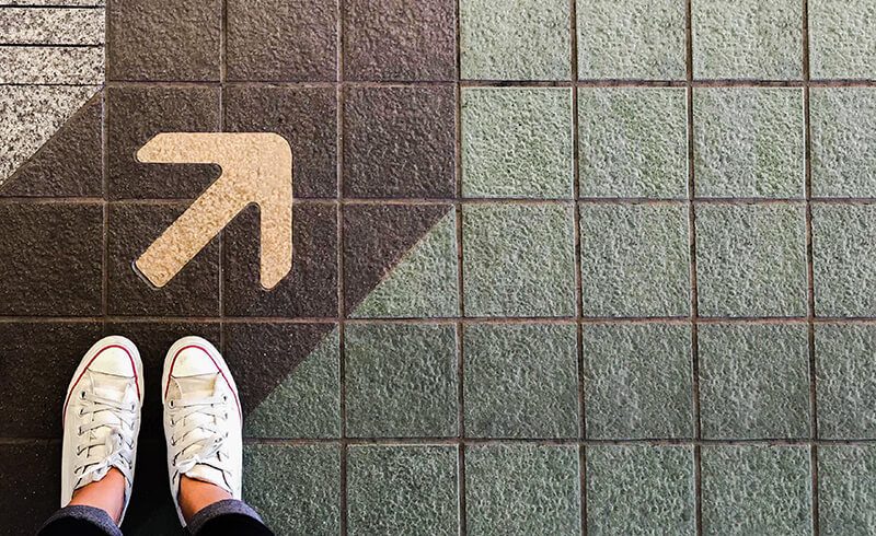 This image shows a birds-eye view of white shoes in the bottom-left corner. Above the feet, painted on the tiled ground is an arrow pointing up to the top-right hand corner - Navigating Your eLearning Feedback Journey - eLearning Program Development - Custom Learning