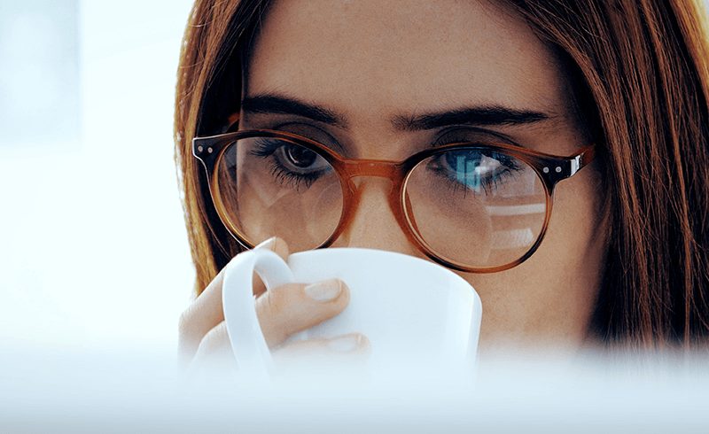 Close-up of a woman's face, with long brown hair and glasses, drinking from a white mug.