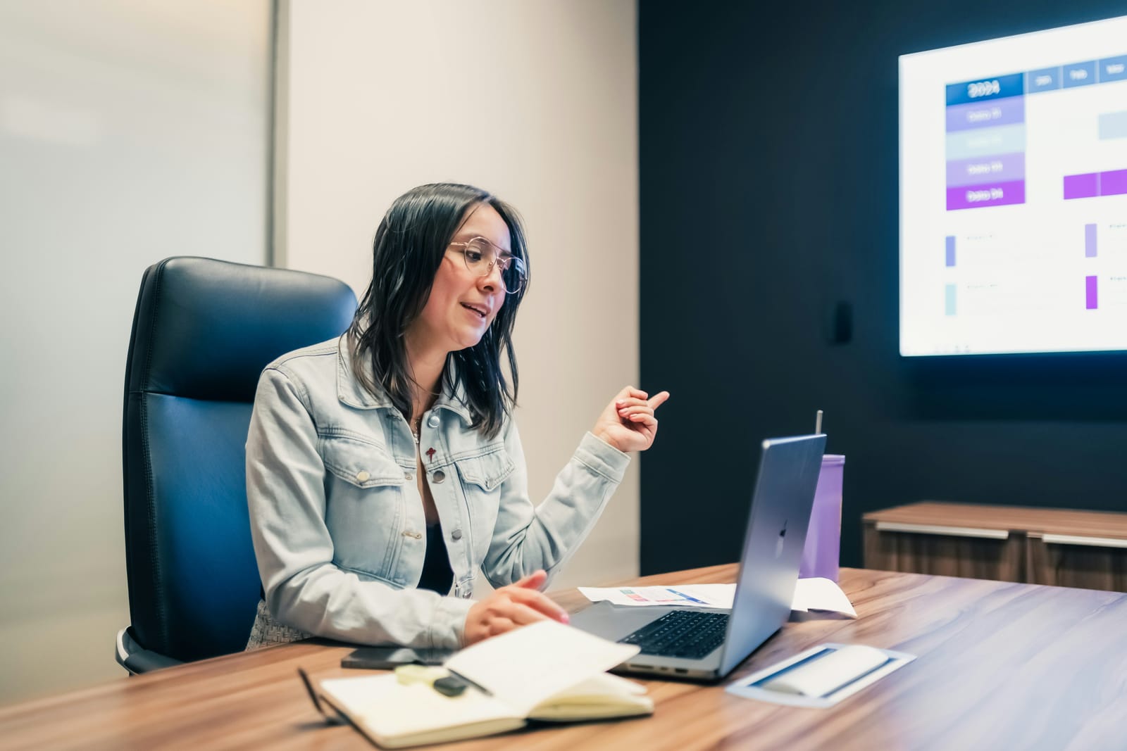 A woman at a desk presenting a dashboard shown on the screen behind her