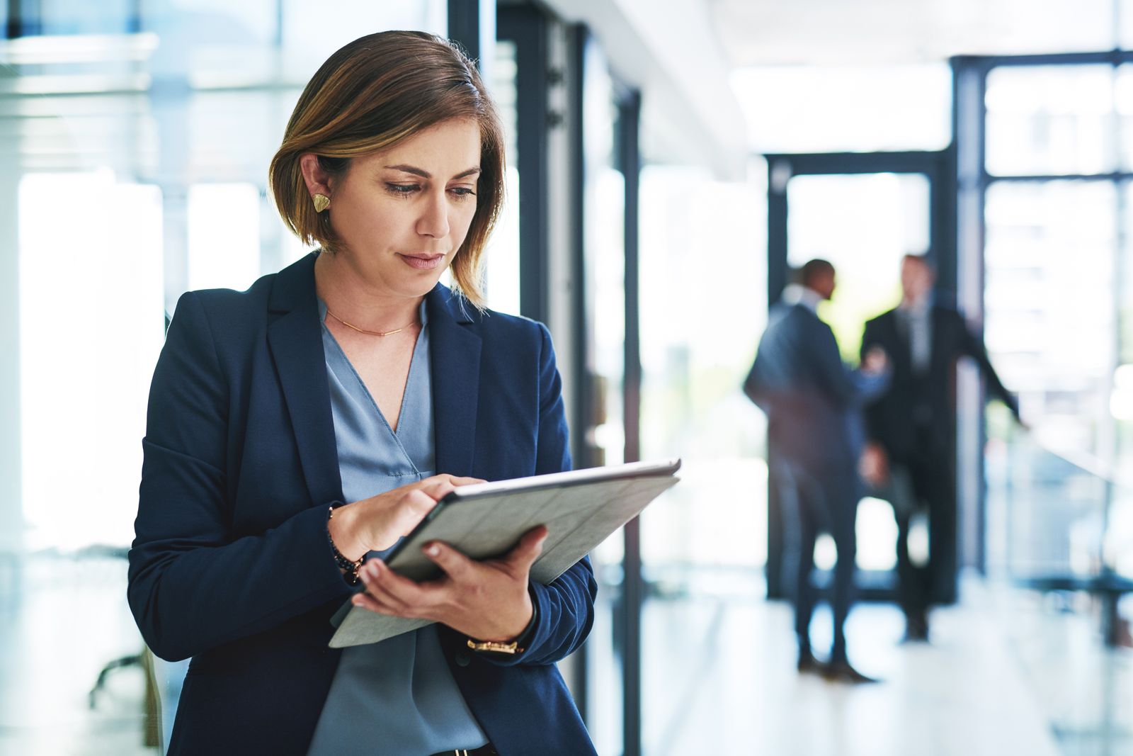 A woman in a blazer reading a tablet in a glass-walled office