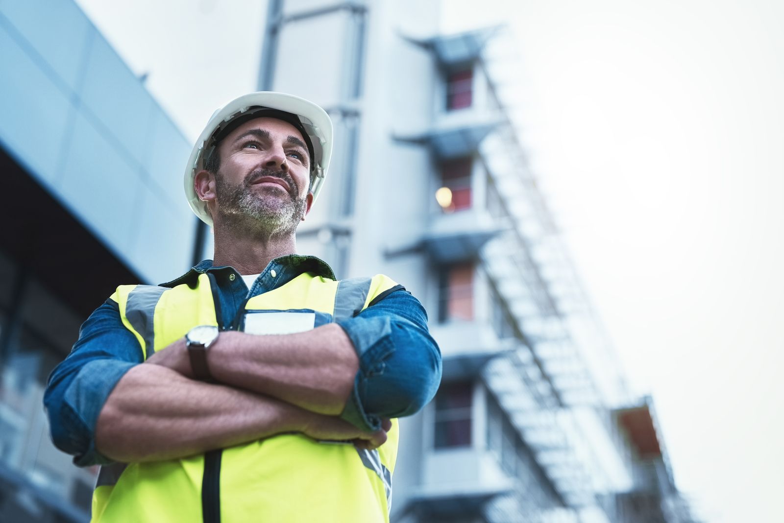 A worker in a hard hat and high-visibility vest, arms crossed, looks up at a building site