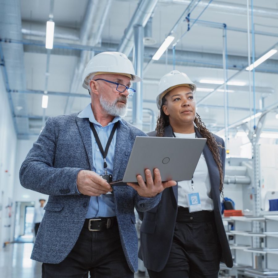 Two managers in hard hats walk a plant floor, one carrying a laptop.