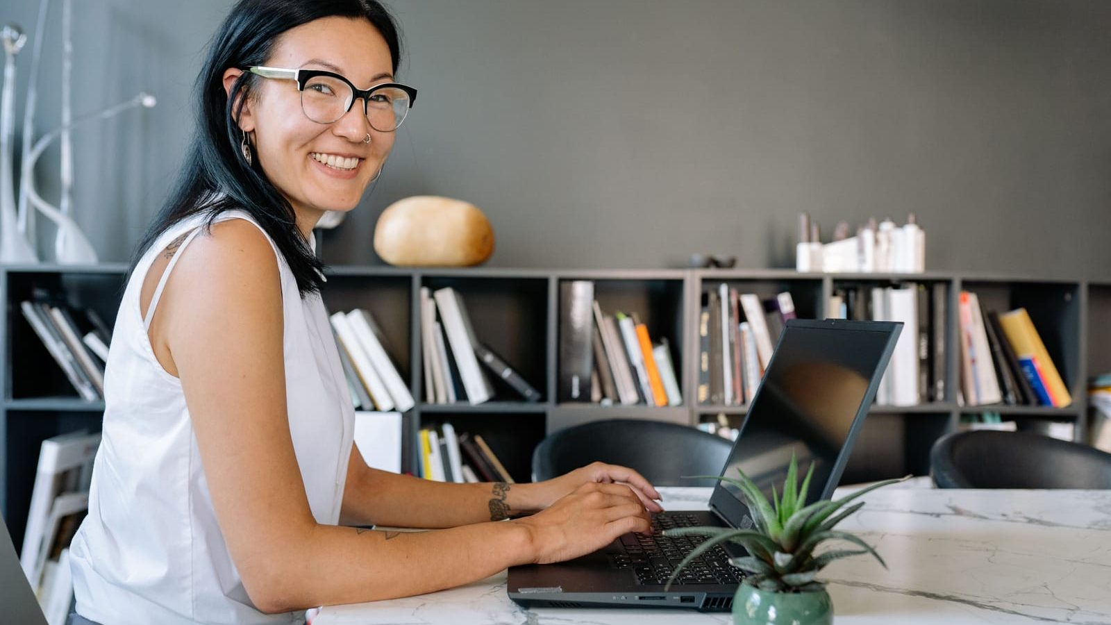 A smiling woman at a laptop in front of a bookshelf
