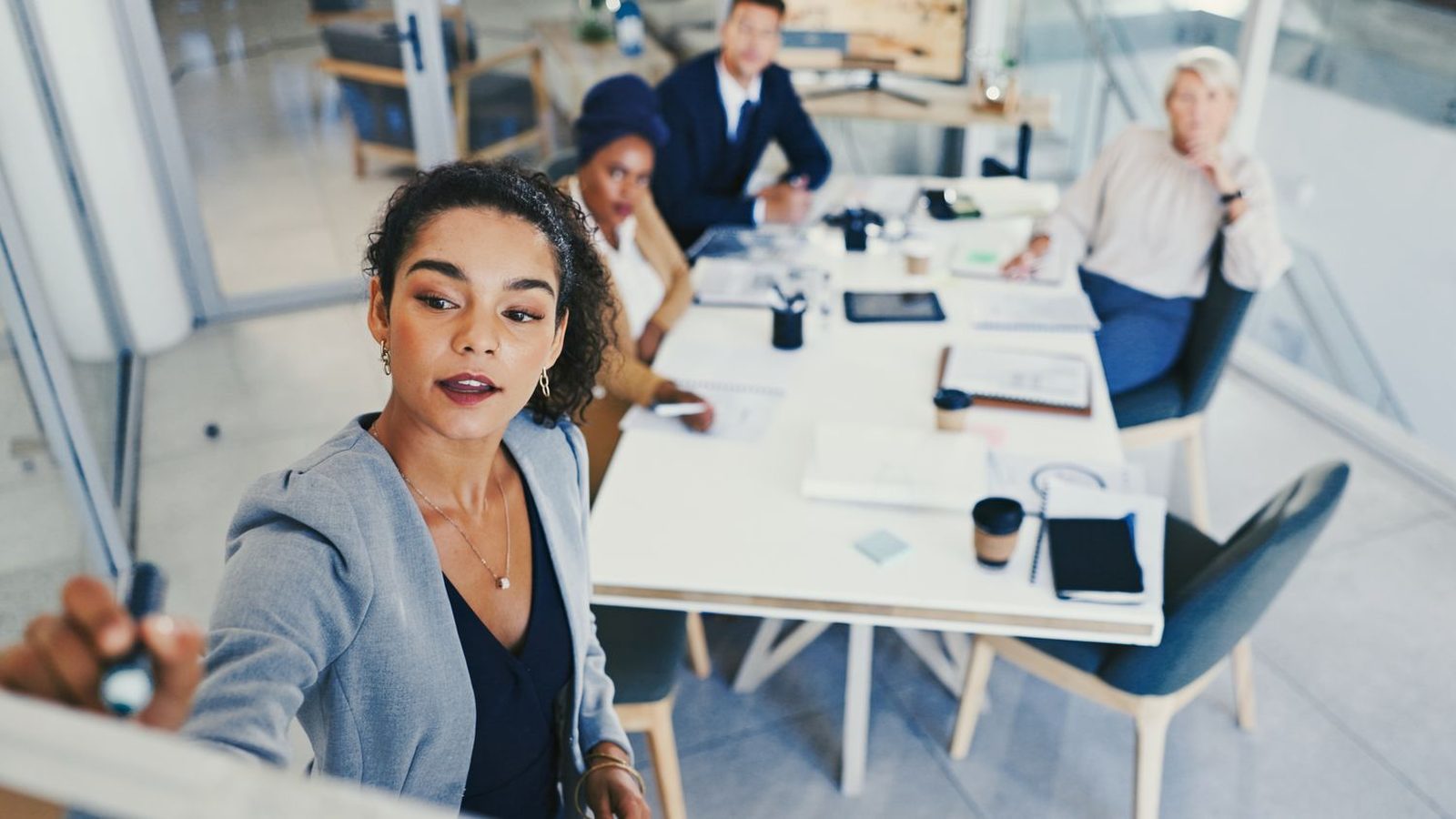 A woman maps out a process on a whiteboard while her team watches from the meeting table.