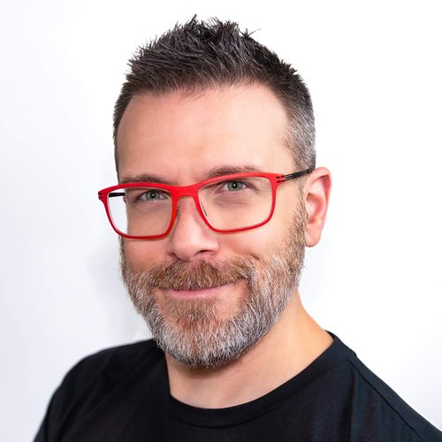 Adam Lamb, smiling, with short hair and bright red glasses