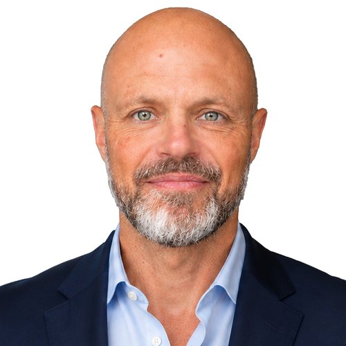 Smiling person with a shaved head and a grey beard, in a navy jacket over a light blue shirt